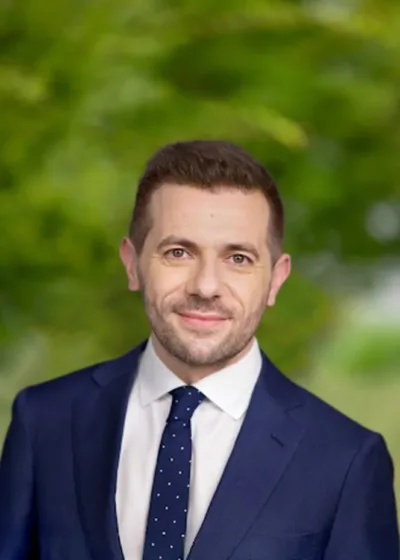 Headshot of Petru Santu Acquaviva, smiling in a navy suit, white shirt, and navy polka-dot tie against a blurred green outdoor background.