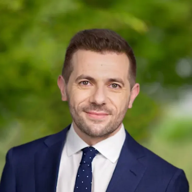 Headshot of Petru Santu Acquaviva, smiling in a navy suit, white shirt, and navy polka-dot tie against a blurred green outdoor background.