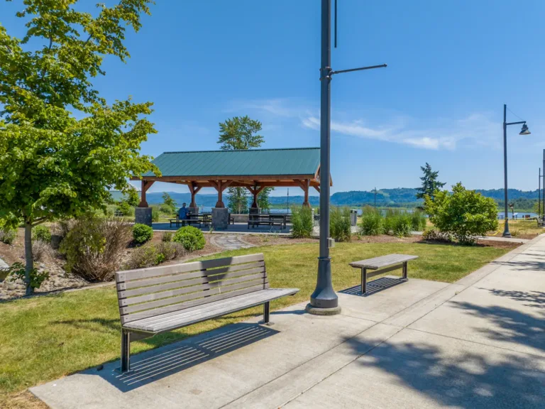Waterfront park featuring a wooden picnic pavilion with a green metal roof, benches and lampposts along a paved walkway, and distant hills under a clear blue sky.