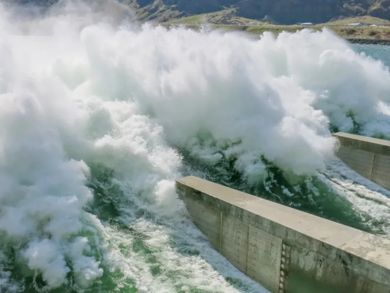Turbulent whitewater surges through a dam spillway, crashing against concrete walls as water is released.