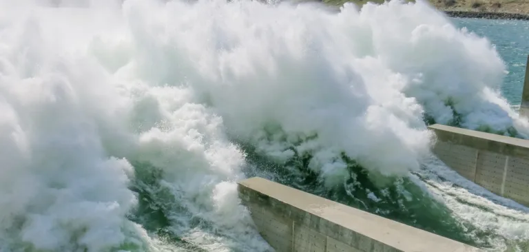 Turbulent whitewater surges through a dam spillway, crashing against concrete walls as water is released.