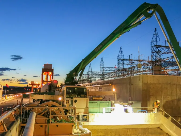 Construction equipment with green booms operates on a dam next to a power substation at dusk, with a worker on a concrete platform and a blue‑orange sky in the background.