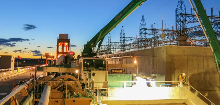 Construction equipment with green booms operates on a dam next to a power substation at dusk, with a worker on a concrete platform and a blue‑orange sky in the background.