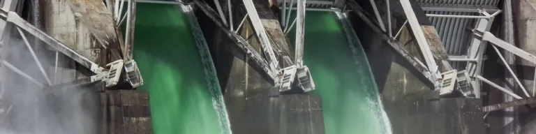 Three open spillway gates of a concrete dam release foaming green water into the turbulent river below.