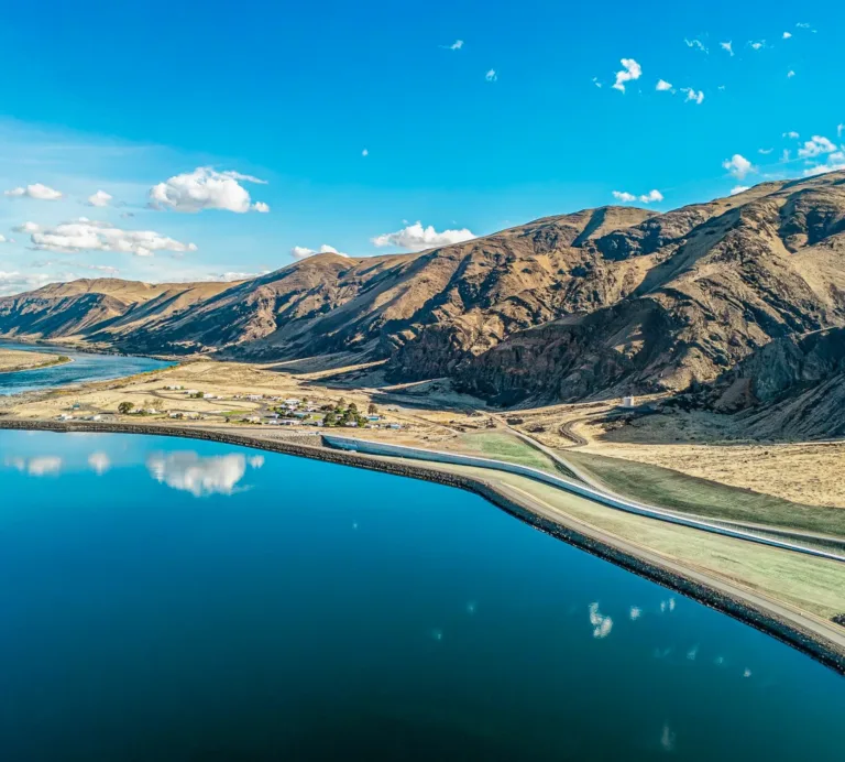 Aerial view of a calm blue reservoir bordered by a long embankment and winding road, with arid mountains and a river valley under a clear, cloud-dotted sky.
