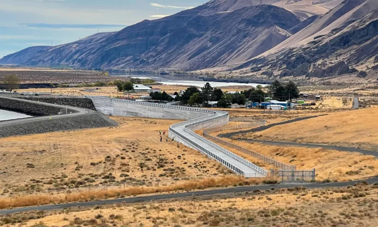 A long, winding fenced walkway crosses a dry golden hillside toward a river and dam, with a few people on the path and rugged brown mountains in the background.