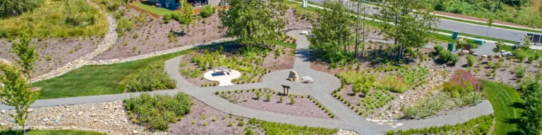 Aerial view of a suburban community park with winding gravel paths, native plant beds, boulders, and a dry creek bed, bordered by green lawns, houses, and a forested backdrop.