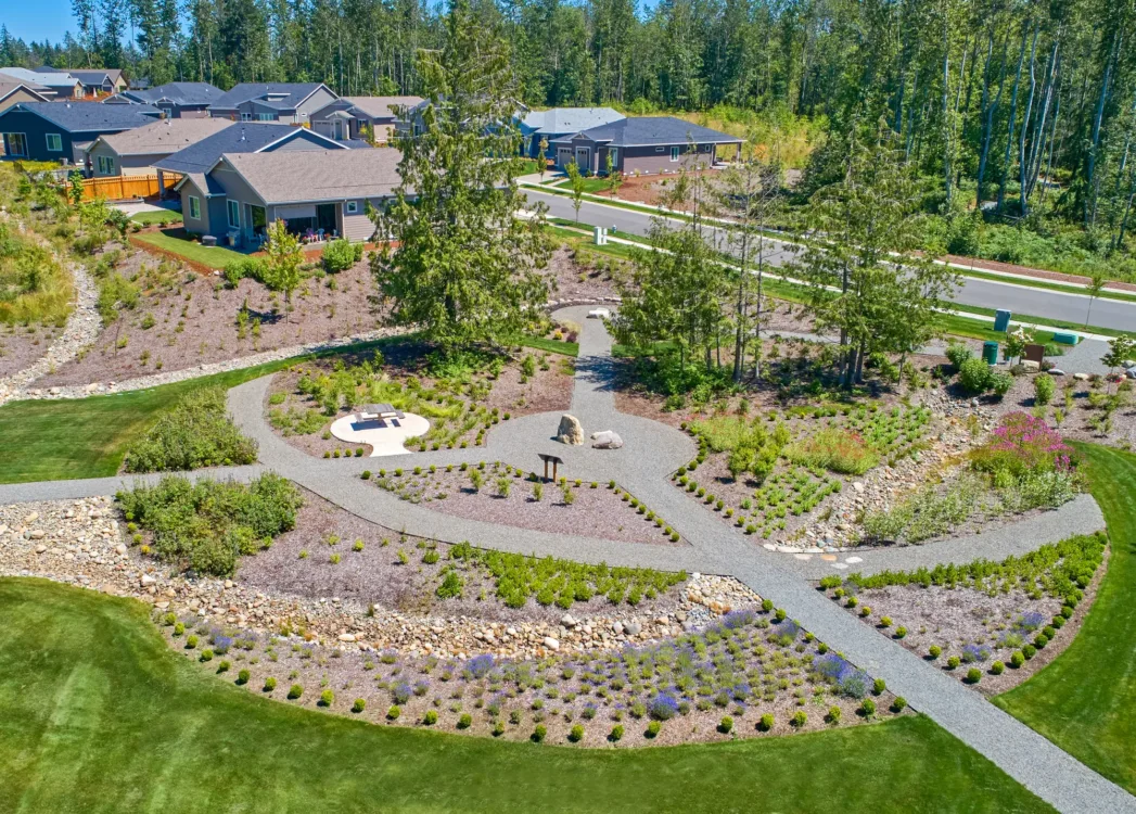 Aerial view of a suburban neighborhood green space with winding gravel paths, native plant beds, small boulders, and a circular picnic area, bordered by manicured lawns and houses near a forested backdrop.