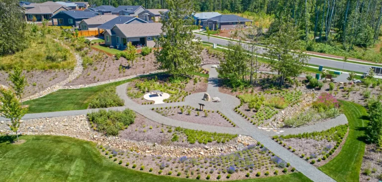 Aerial view of a suburban neighborhood park with curving gravel paths, a dry creek bed, young shrubs and boulders, a small picnic area, and manicured lawns bordered by single-story homes and forest.
