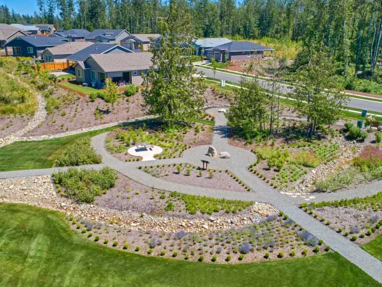 Aerial view of a landscaped neighborhood park with winding gravel paths, a circular picnic table area, manicured lawns, and surrounding houses and trees on a sunny day.