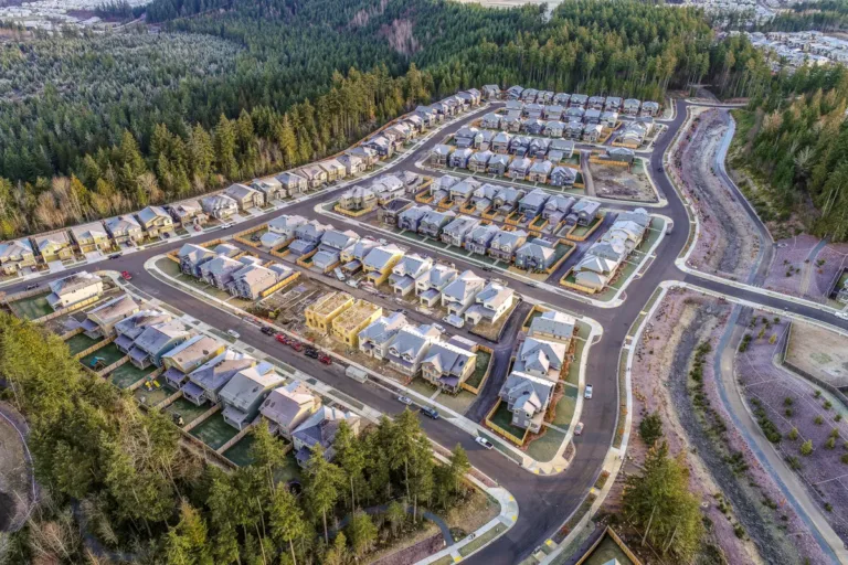 Aerial view of a new suburban housing development with rows of gray‑roofed homes, several houses under construction, curving streets, and forested hills surrounding the neighborhood.