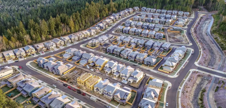 Aerial view of a new suburban neighborhood under construction, with rows of similar houses, curved streets and fenced yards, a drainage canal on the right, and dense forest surrounding the development.