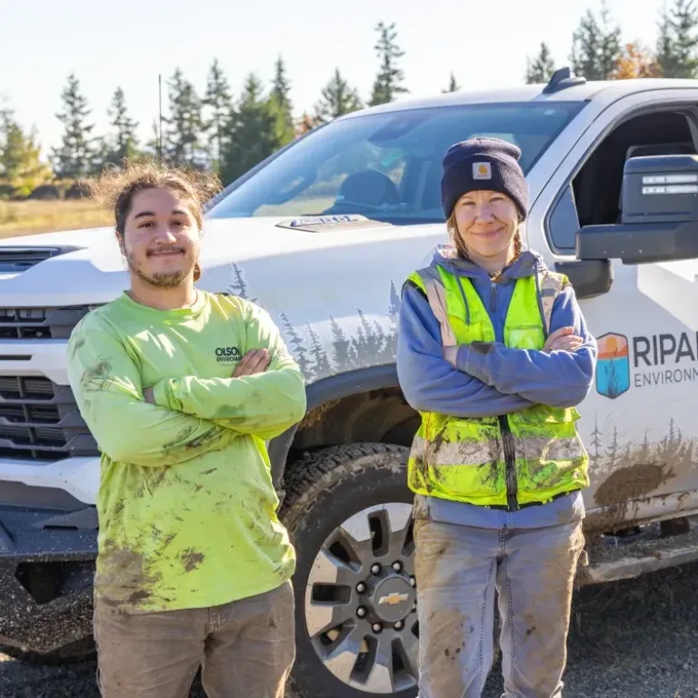MacKay Sposito field crew members stand with arms crossed beside a mud-splattered pickup truck, wearing high-visibility safety gear at an outdoor job site.