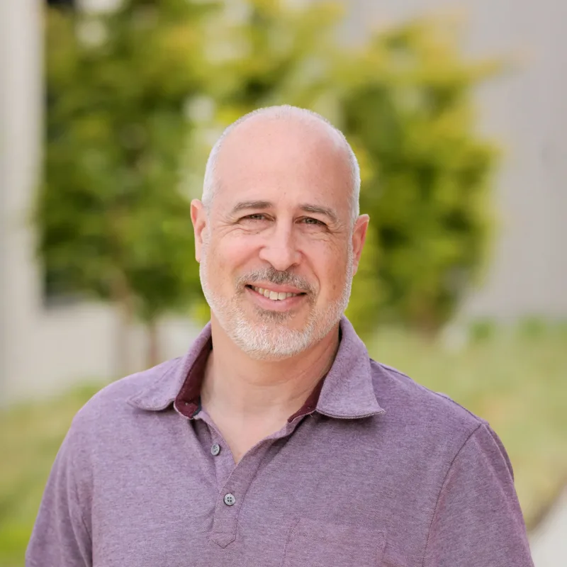 Headshot of Rob Palena, a smiling man with a shaved head and short gray beard wearing a light purple polo, outdoors with blurred greenery in the background.