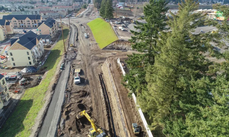 Aerial view of a suburban road construction site with excavators and trucks shaping a gravel roadbed and retaining wall between new apartment buildings and tall evergreen trees.
