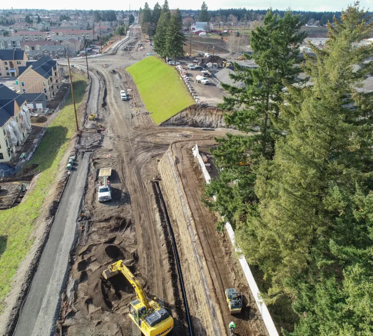 Aerial view of a suburban road construction site with excavators and trucks shaping a gravel roadbed and retaining wall between new apartment buildings and tall evergreen trees.