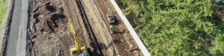 Aerial view of a road construction site with excavators, trucks, and workers grading a long terraced embankment and gravel corridor beside a white barrier and dense evergreen trees.