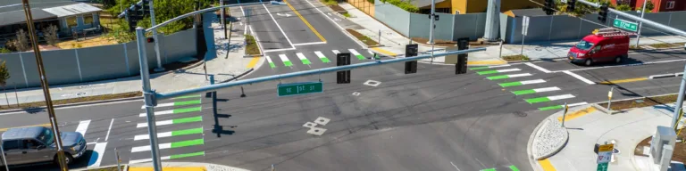 Aerial view of a signalized suburban intersection with green-striped bike crossings and white crosswalks, curb ramps and sidewalks on each corner, and a few cars passing near houses and power lines.