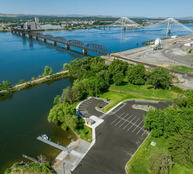 Aerial view of a riverside park and boat launch with an empty parking lot and dock beside a narrow lagoon, opening to a wide river spanned by an old arched truss railroad bridge and a modern cable-stayed bridge on a clear day.