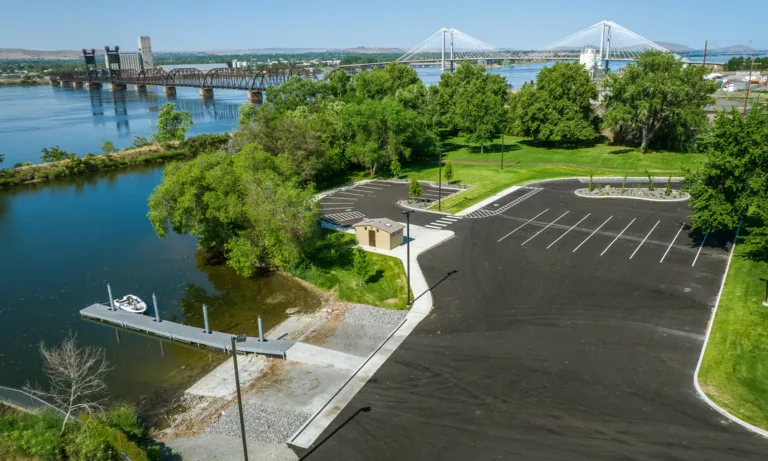 Aerial view of a riverside boat launch with a dock and empty parking lot bordered by green trees, with two large bridges spanning the river in the background under a clear sky.