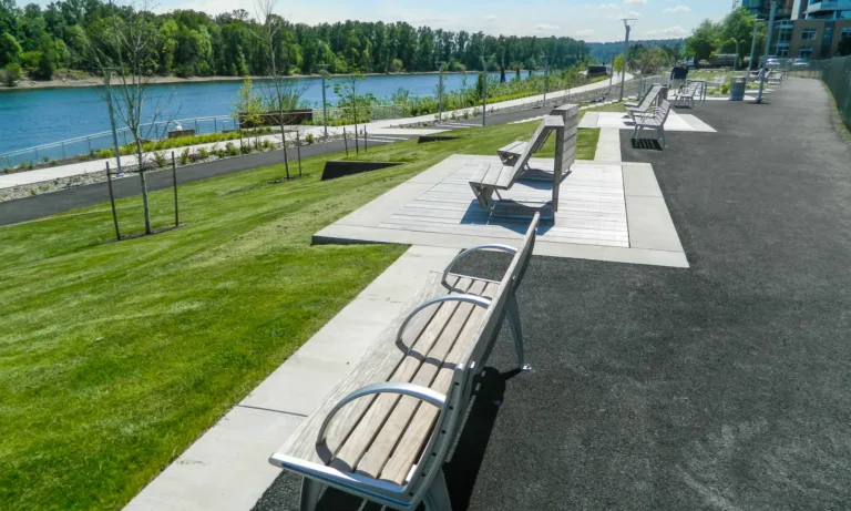 Benches on wooden pads line a paved riverside promenade with manicured lawns, young trees, and railings along a calm river, with modern high-rise buildings in the background under a clear blue sky.