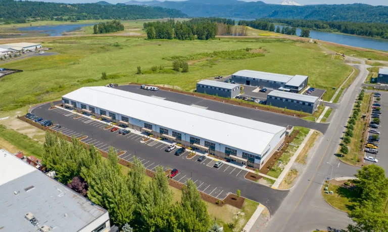 Aerial view of a modern industrial park with long white‑roofed warehouse buildings and parking lots, surrounded by green fields near a river with forested hills and a distant snow-capped mountain under a clear blue sky.