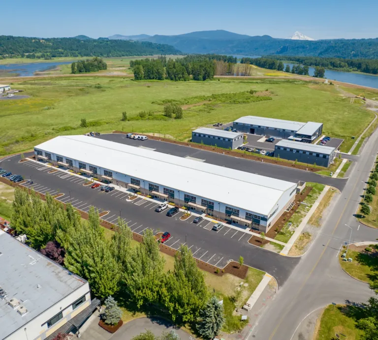 Aerial view of a modern industrial park with long white‑roofed warehouse buildings and parking lots, surrounded by green fields near a river with forested hills and a distant snow-capped mountain under a clear blue sky.