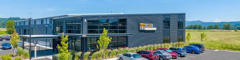 Modern two-story commercial building with dark gray metal siding and large glass windows, a landscaped parking lot with cars, and open fields with mountains in the background under a clear blue sky.