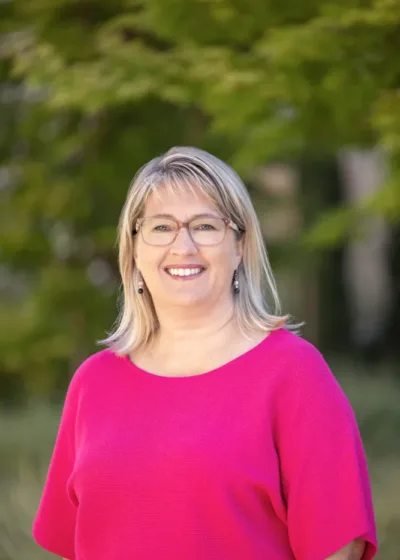 Headshot of Stephanie Cameron smiling, wearing glasses and a bright pink top, with a soft-focus green leafy background.