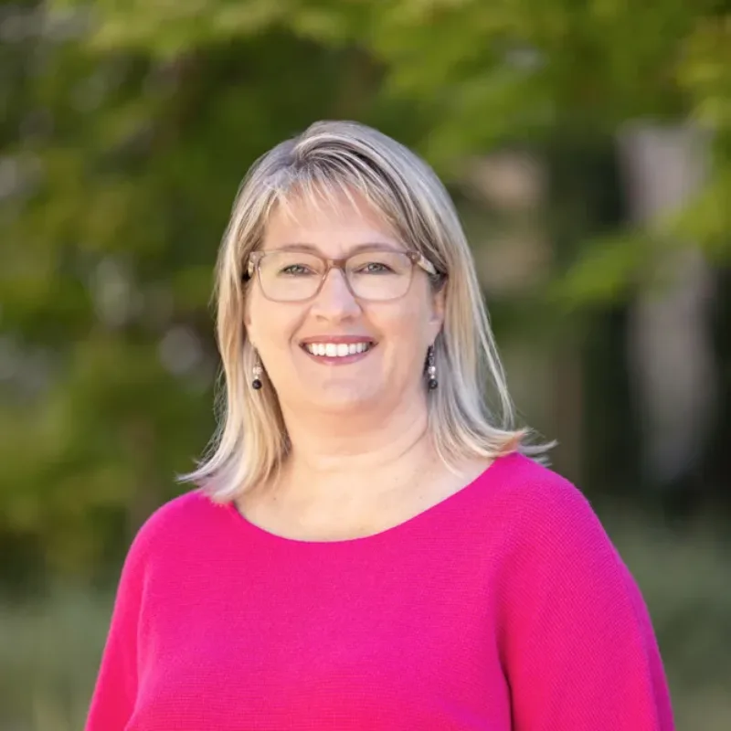 Headshot of Stephanie Cameron smiling, wearing glasses and a bright pink top, with a soft-focus green leafy background.