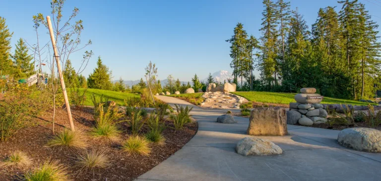 Curving paved pathway through a landscaped park with boulders, native grasses, and evergreen trees, featuring a stacked cairn labeled Story Stone and a distant snow-capped mountain under a clear blue sky.