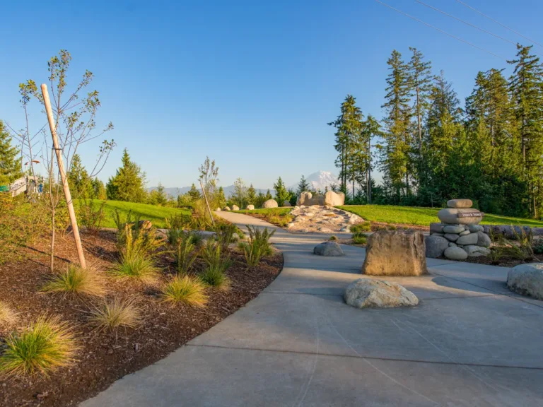Curving paved path through a landscaped park with boulders and a rock cairn labeled “Sticks N’ Stones,” surrounded by evergreen trees under a clear blue sky, with a snow-capped mountain in the distance.