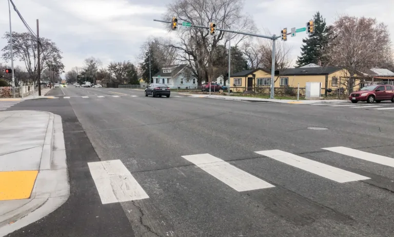 Wide residential intersection with striped crosswalks, a yellow tactile curb ramp, and traffic lights as a few cars pass under a cloudy sky.