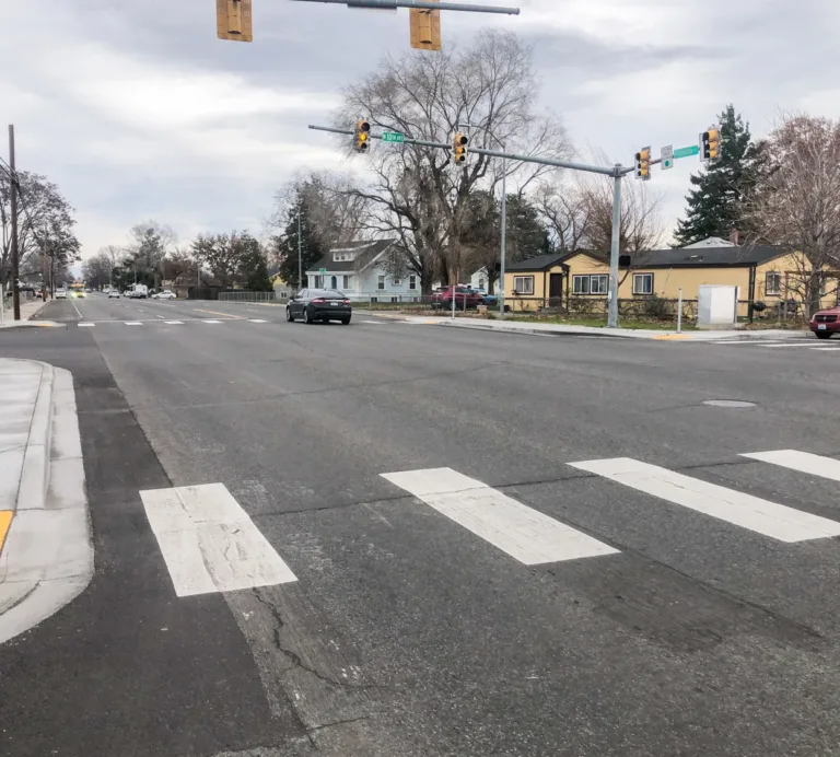 Wide residential intersection with striped crosswalks, a yellow tactile curb ramp, and traffic lights as a few cars pass under a cloudy sky.