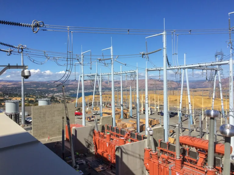 High‑voltage electrical substation with steel frameworks, insulators, transformers and overhead wires, with equipment crates and service trucks below and arid hills and a river in the background under a clear blue sky.