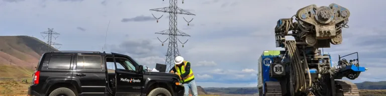 Field engineer in a high-visibility vest stands beside a MacKay Sposito SUV, taking notes next to a large tracked drilling rig at a remote site with power transmission towers and hills under a cloudy sky.