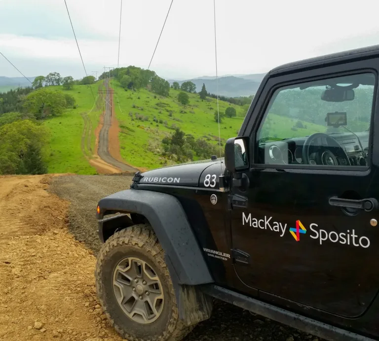 A black Jeep Wrangler with “MacKay Sposito” branding is parked on a muddy ridge beside a utility access road, with power lines stretching across rolling green hills under an overcast sky.
