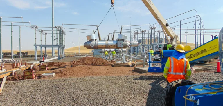 Workers in high-visibility gear guide a crane lifting large switchgear at an electrical substation construction site, with wind turbines on distant hills.