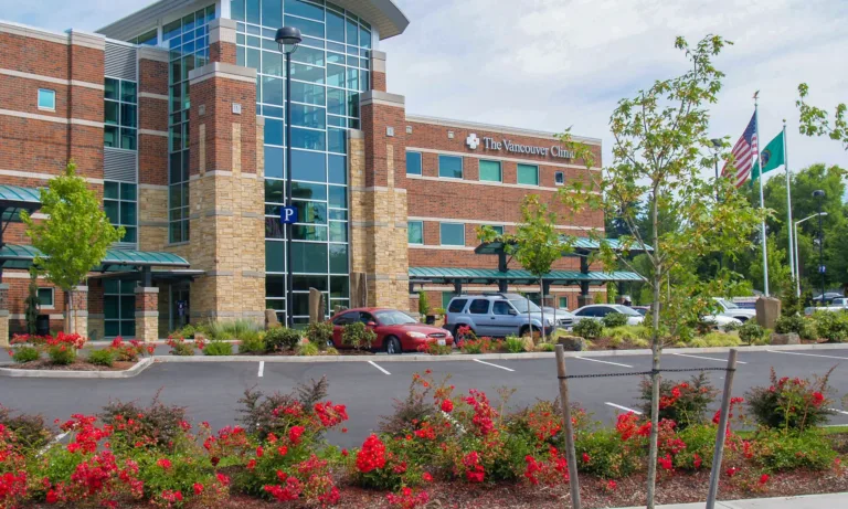 The Vancouver Clinic’s modern brick-and-glass building with a central glass atrium, a front parking lot with cars, landscaped red flowers and young trees, and U.S. and Washington state flags on poles.