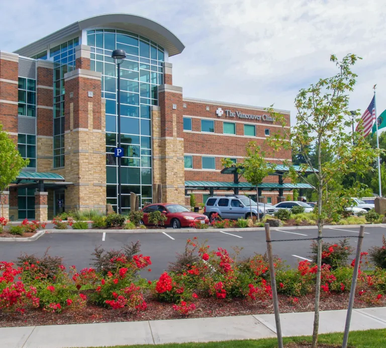 The Vancouver Clinic’s modern brick-and-glass building with a central glass atrium, a front parking lot with cars, landscaped red flowers and young trees, and U.S. and Washington state flags on poles.