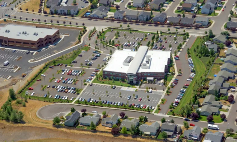 Aerial view of a large brick-and-glass building with a white roof and central curved entrance, surrounded by landscaped parking lots filled with cars and bordered by suburban homes and streets.