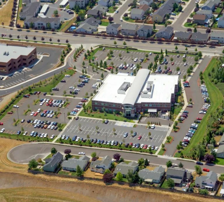 Aerial view of a large brick-and-glass building with a white roof and central curved entrance, surrounded by landscaped parking lots filled with cars and bordered by suburban homes and streets.