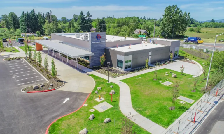Aerial view of a modern, single-story Vancouver Clinic building with a covered drop-off, empty parking lot, and landscaped lawns set against trees and open fields.