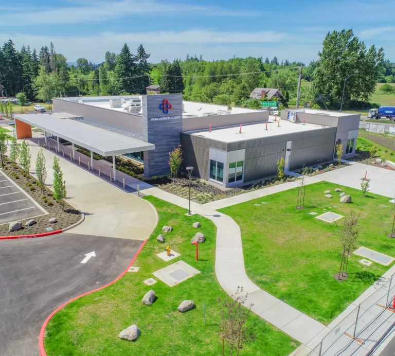 Aerial view of a modern, single-story Vancouver Clinic building with a covered drop-off, empty parking lot, and landscaped lawns set against trees and open fields.