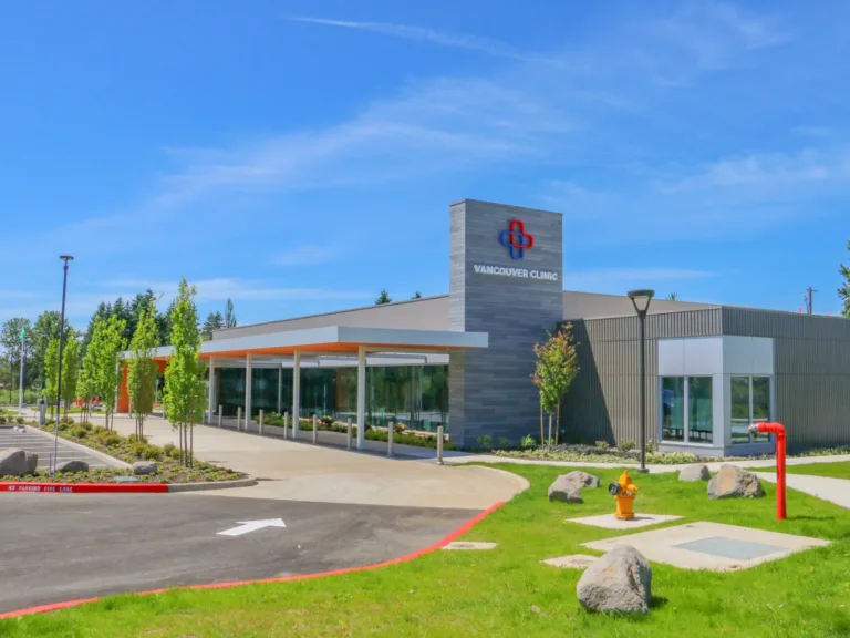 Modern single-story medical clinic with a glass-front entrance and covered drop-off, a sign tower reading “VANCOUVER CLINIC,” landscaped trees, and a sunny blue sky.