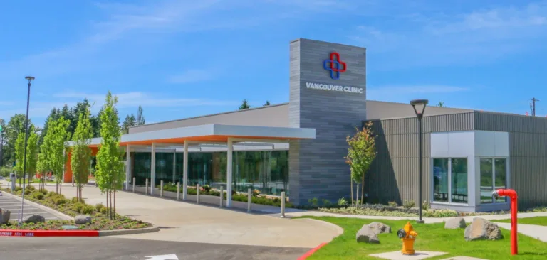 Modern single-story medical clinic with a glass-front entrance and covered drop-off, a sign tower reading “VANCOUVER CLINIC,” landscaped trees, and a sunny blue sky.