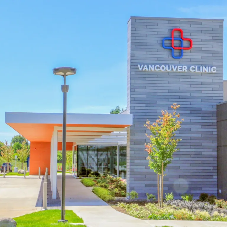 Modern Vancouver Clinic building with a red-and-blue cross logo, gray brick facade, and a covered entrance amid landscaped grounds under a clear blue sky.