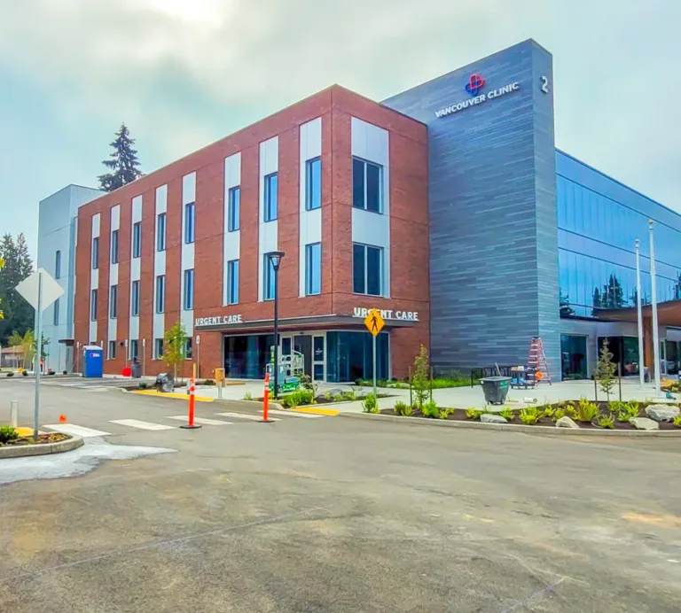 The Vancouver Clinic building with an Urgent Care entrance, featuring a brick-and-glass facade and a mostly empty parking lot with young trees on an overcast day.