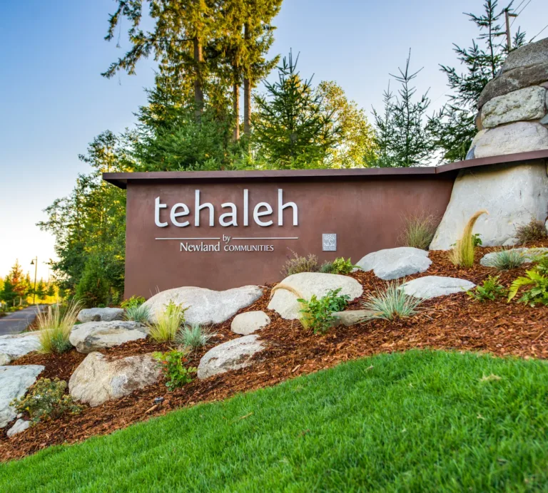Entrance monument sign reading “tehaleh by Newland Communities,” surrounded by landscaped rocks and greenery with evergreen trees in the background.