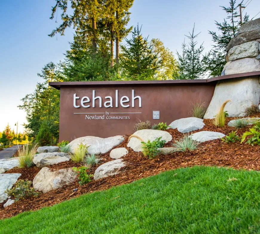 Entrance monument sign reading “tehaleh by Newland Communities,” surrounded by landscaped rocks and greenery with evergreen trees in the background.